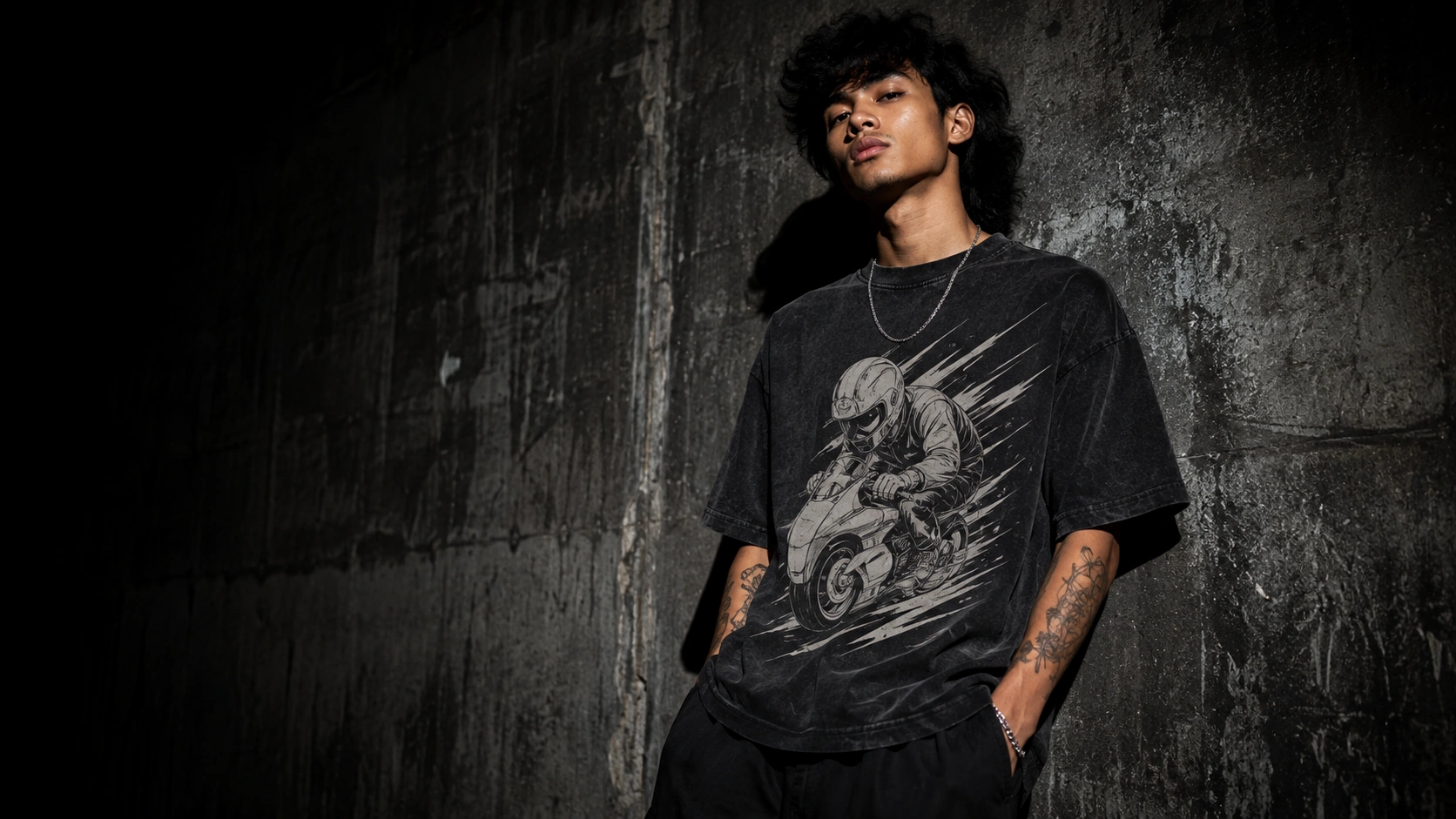 Young man in a black motorcycle graphic T-shirt leaning against a concrete wall